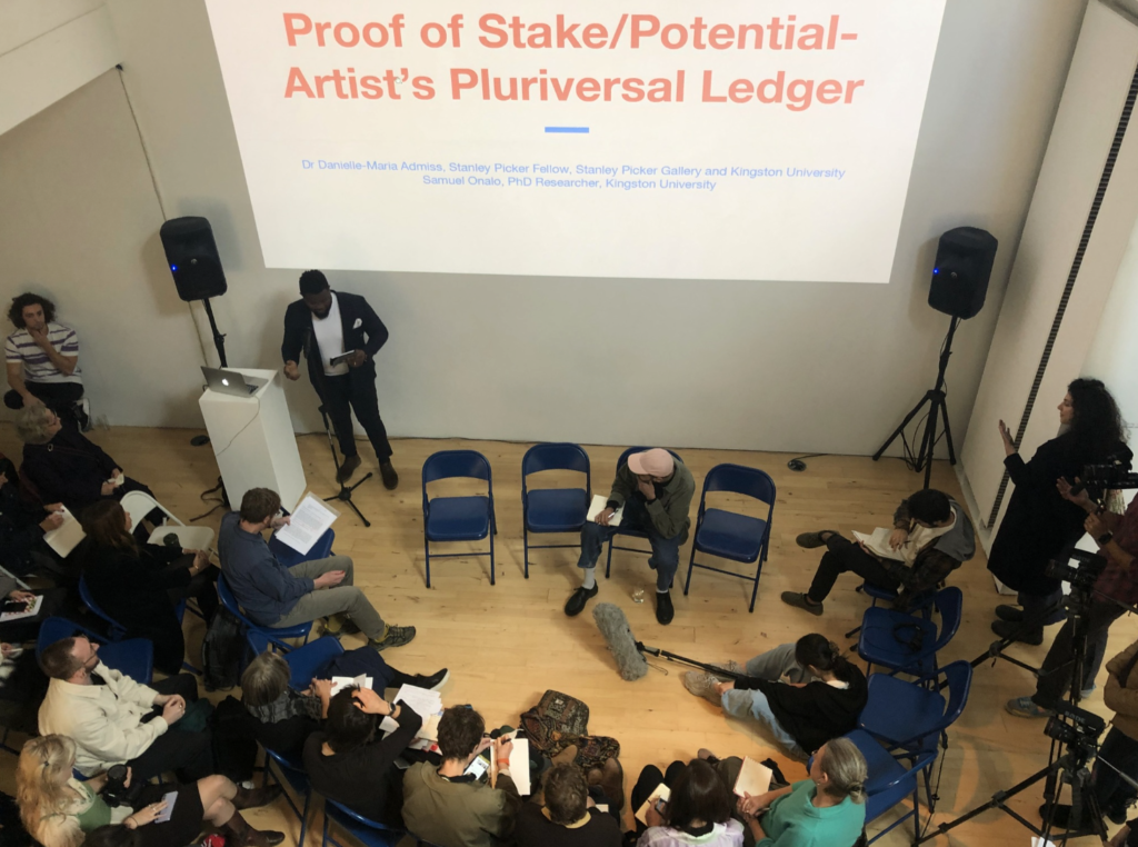 A man stands and presents to the audience who are sat in a semicircle on blue chairs. On the screen reads Proof of Stake/Potential/Artist's Pluriversal Ledger.