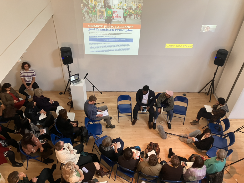 People sit in a semi circle on blue chairs in a gallery. On the wall is projected Anne Pasek and a slide detailing Climate Justice Alliance.