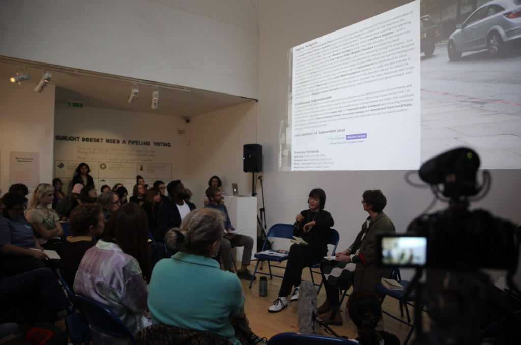Two people sit at the front of the gallery and present to the audience who are sat in a semicircle on blue chairs. On the screen shows a screenshot from an artists website.