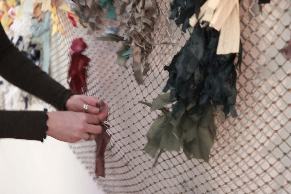 A hand ties a piece of fabric to a fabric fishing net that is hanging on a white wall.