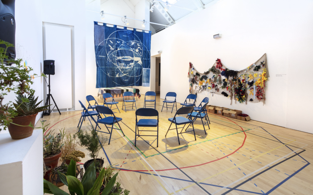 A ring of blue chairs in a white gallery surrounded by plants, a blue wall hanging with white patterns and a fabric fish net installation.
