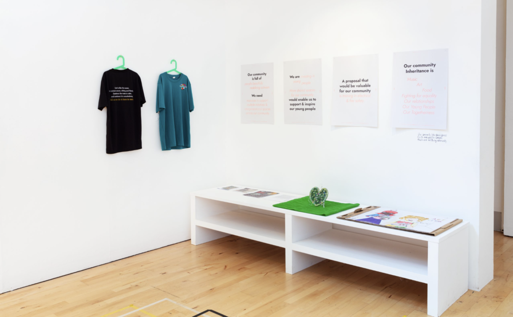 Two tshirts hanging on the wall next to 4 posters with black and pink text. A series of objects sit on a long low table.