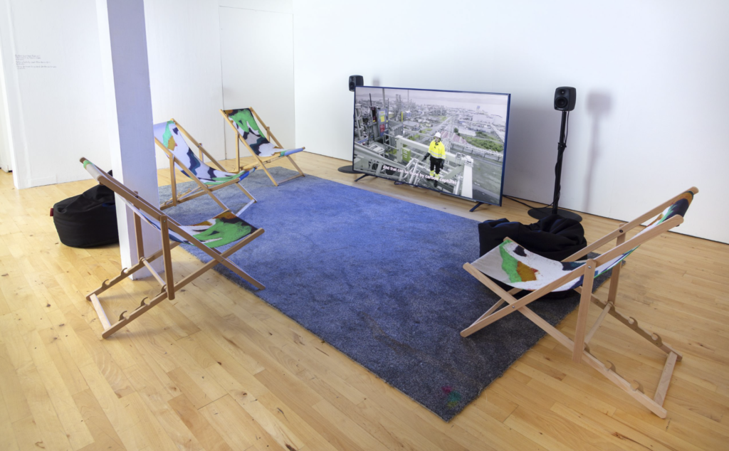 Four coloured deckchairs are placed by a grey square of carpet in front of a large screen TV with a man talking to the camera.
