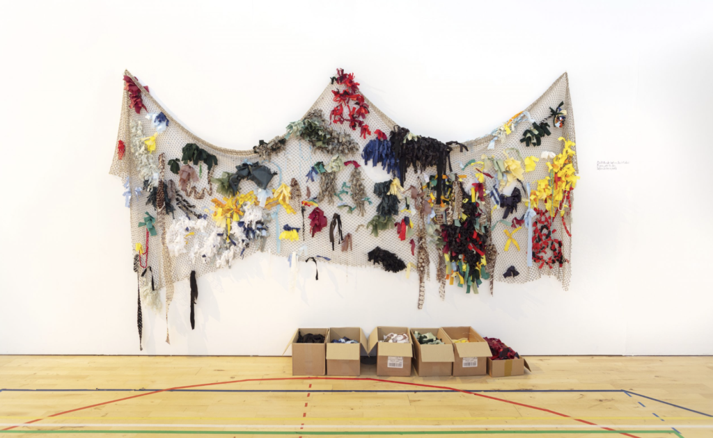 Fabric fishing net hangs on a white wall with strips of coloured fabric tied into it. On the floor sits a series of cardboard boxes with coloured fabric ties in.