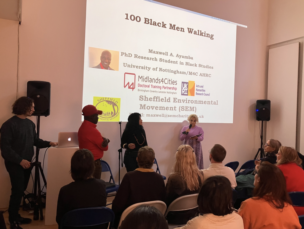 People sit in a semi circle on blue chairs in a gallery. On the wall is projected a slide with the title 100 Black Men Walking, Maxwell A. Ayamba PhD Research Student in Black Studies University of Nottingham/M4C AHRC.