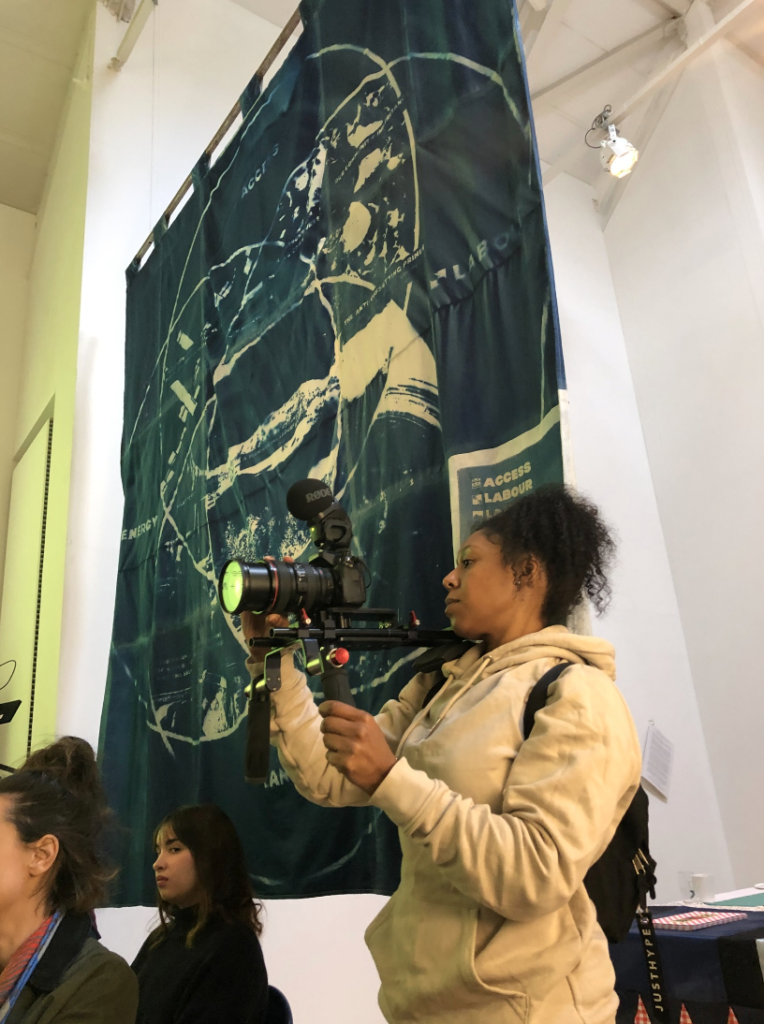 A person holding a professional video camera and rig stands in front of a blue wall hanging with white patterns on.