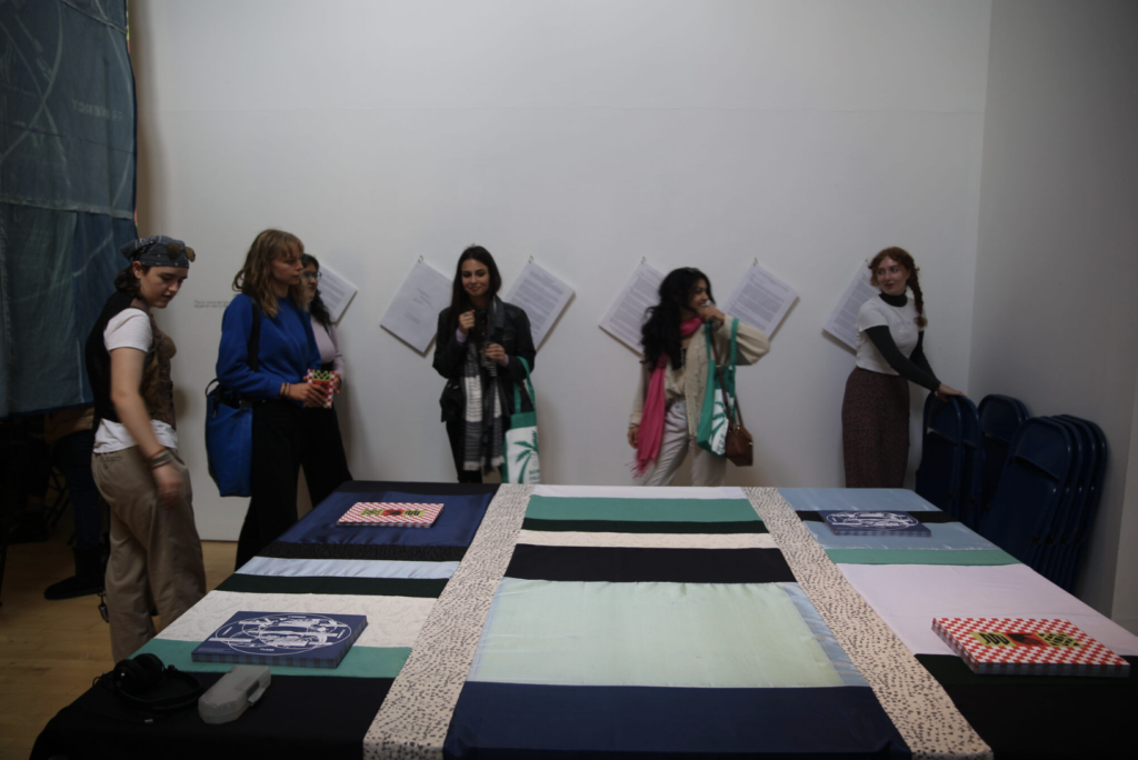 Six people stand around a table in a white gallery. The table has a multiple colour tablecloth on it and a series of posters.