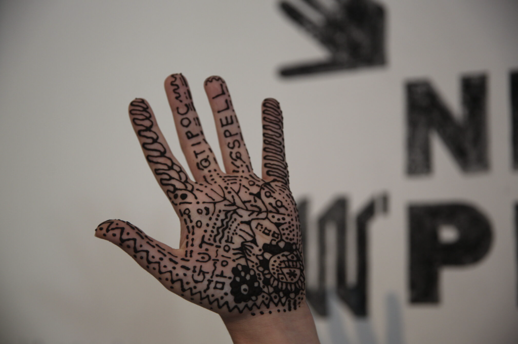 A close up of a hand having had patterns, dots, lines and the words QTIPOC and SPELL drawn on them using Henna.