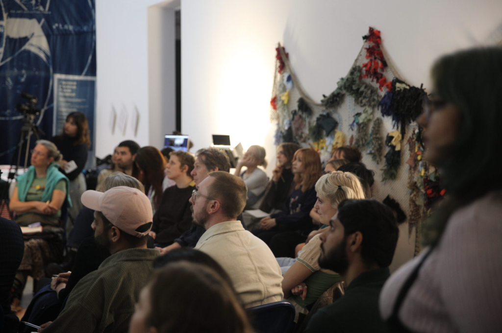 A group of people stand in a gallery. They are blurred in the background hangs [Dis]Entangled Map (2022) fabric fishing net.