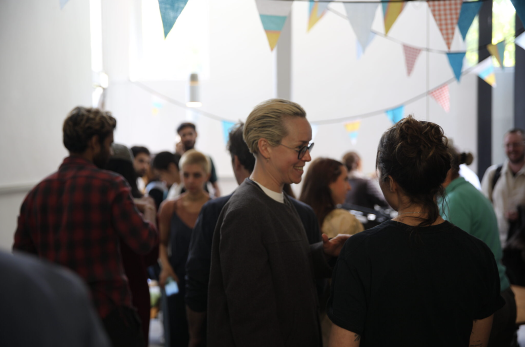 A group of people stand in a gallery. They are blurred in the background hangs coloured bunting.