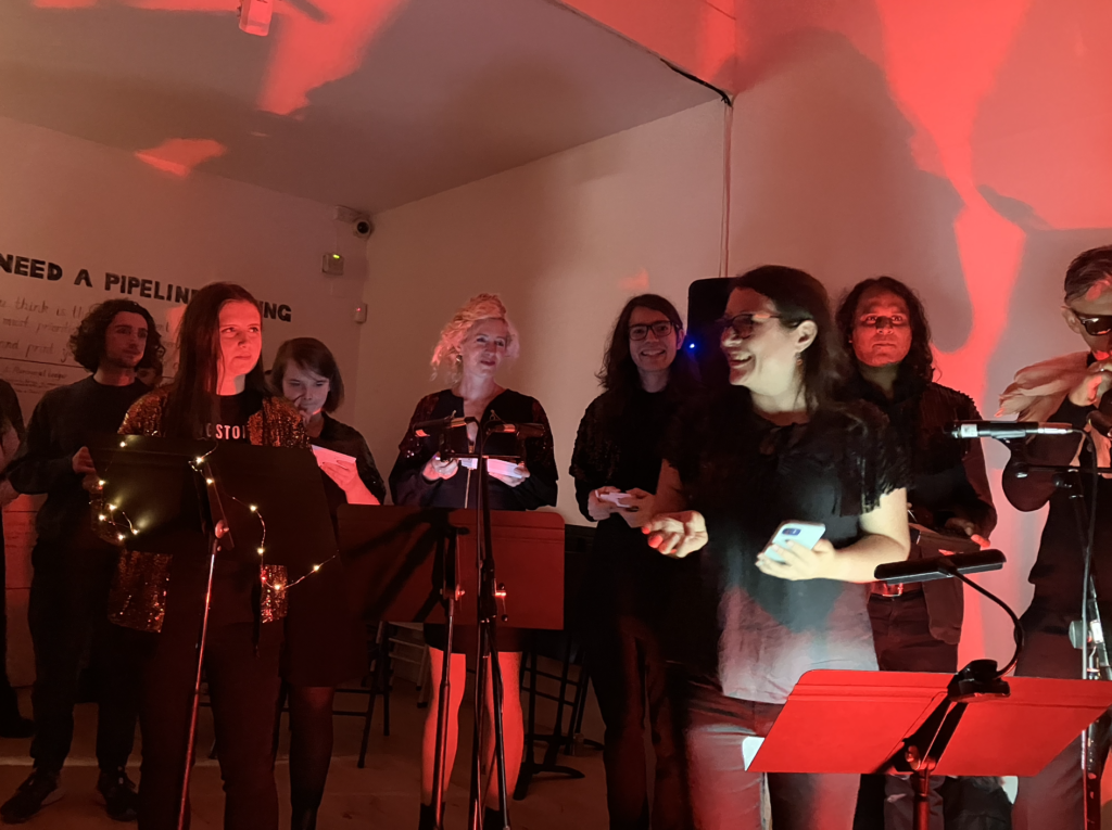 8 members of Kingston Stylophone Orchestra dressed in black play their stylophones in a gallery bathed in warm red light.